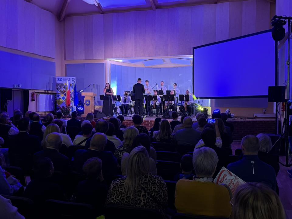 Pomieszczenie wypełnione siedzącymi osobami, w oddali na scenie grająca orkiestra i solistka