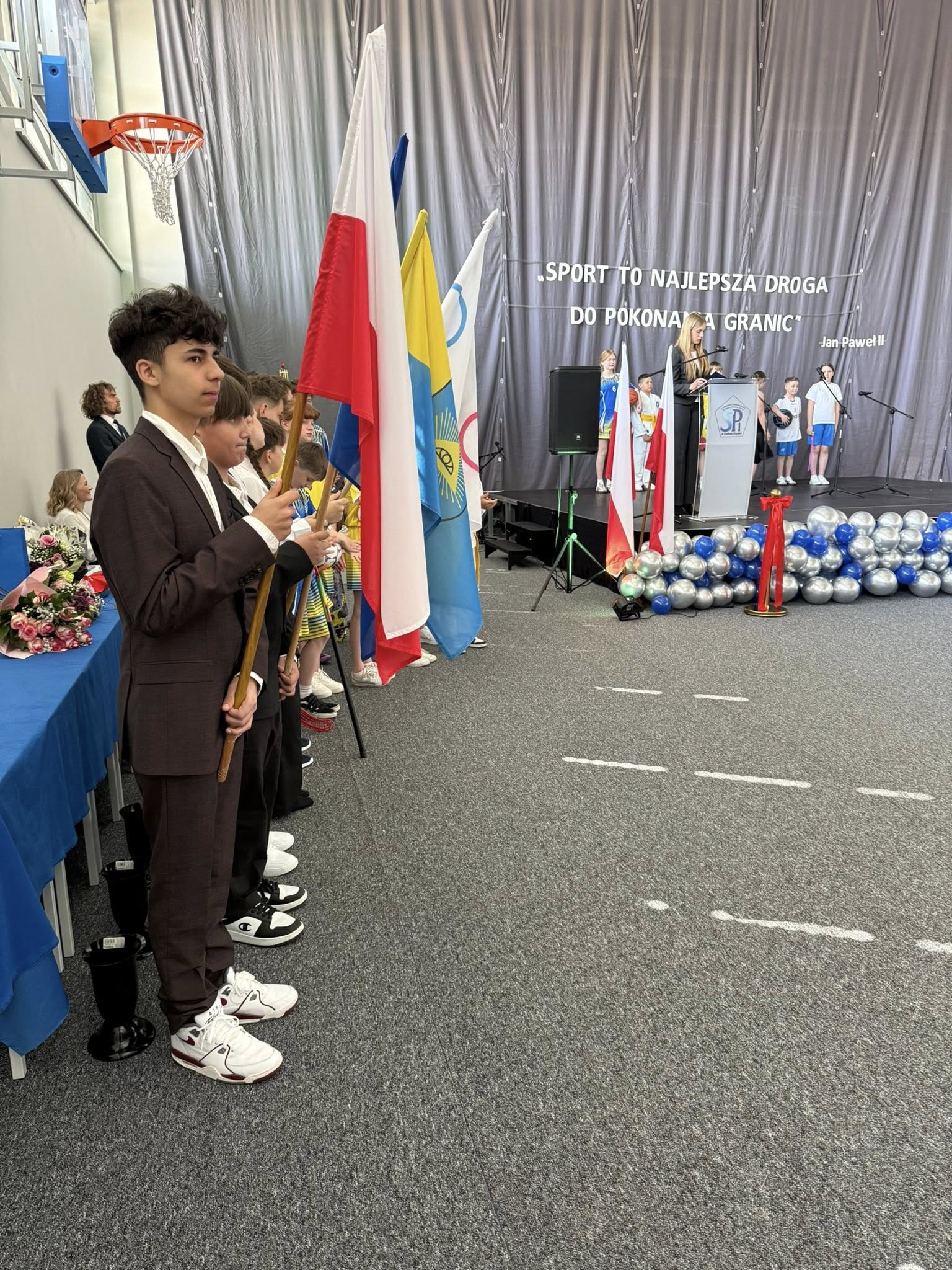 Młodzież stojąca w pomieszczeniu. W dłoniach trzymają flagi 