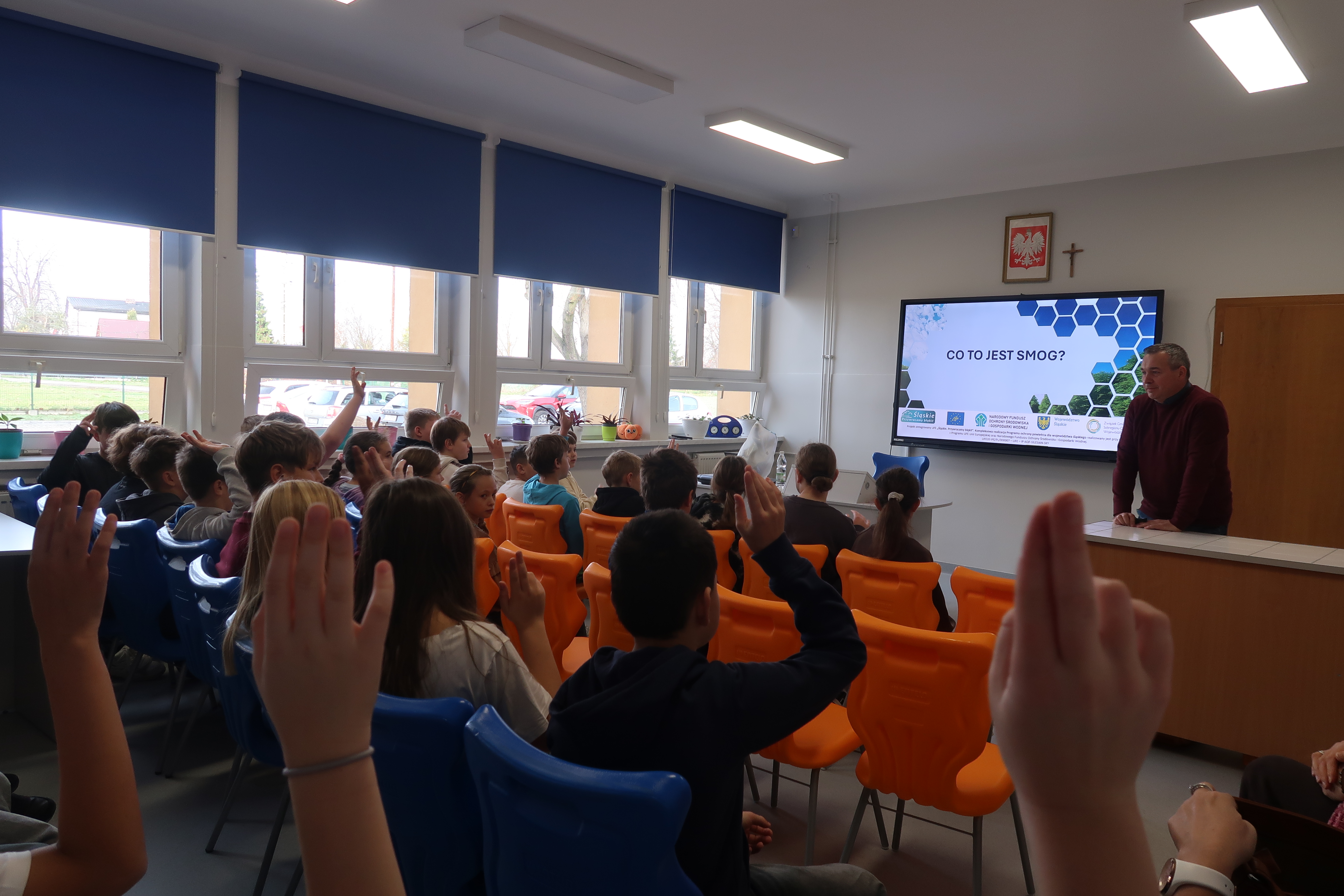 Klas wypełniona uczniami siedzącymi na krzesłąch przed monitorem. Za stołem mężczyzna stojący. 