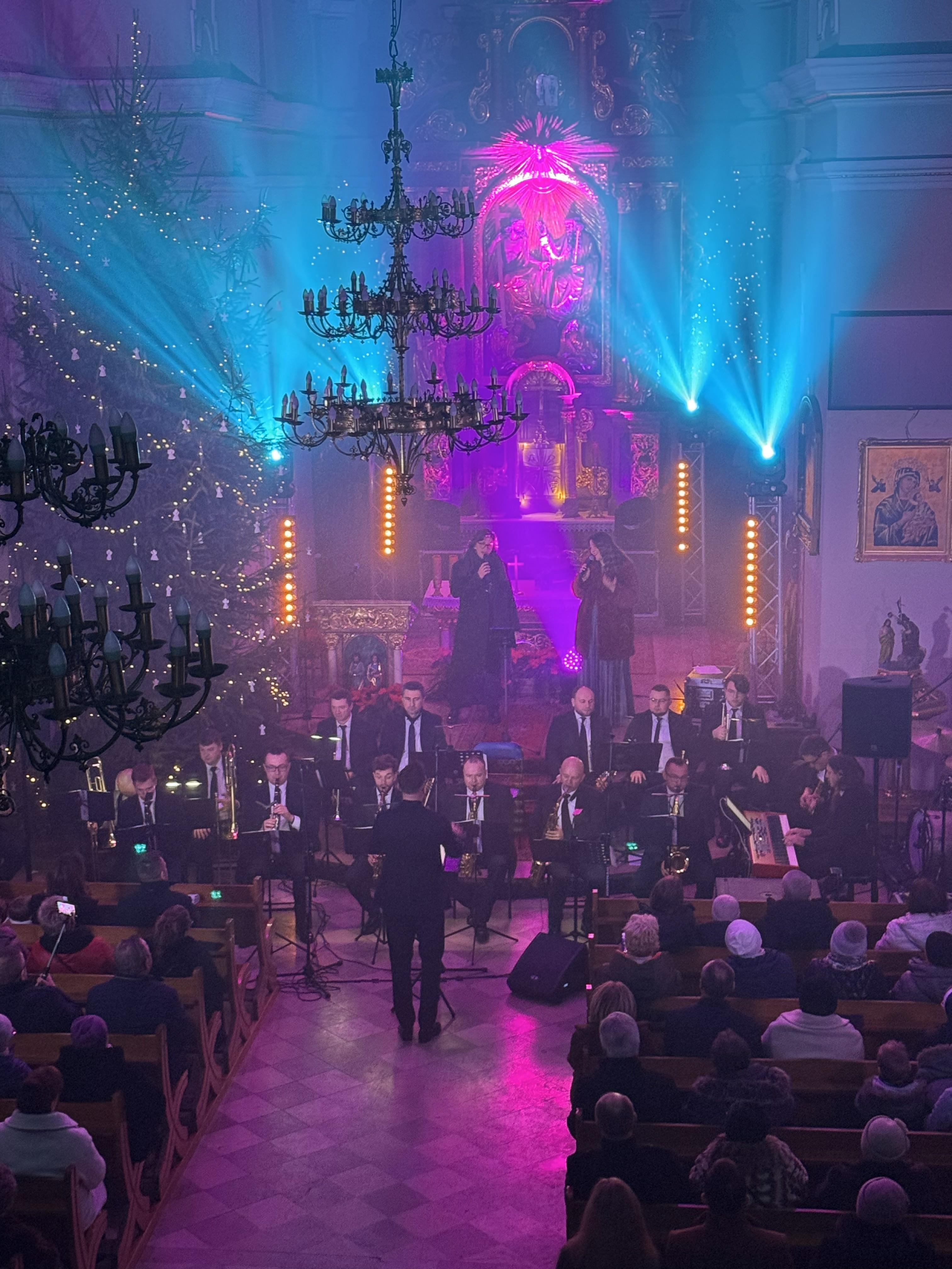 Wnętrze kościoła oświetlone kolorowym światłwem , przed ołtarzem zasiadający muzycy i śpiewający wokaliści. Wierni siedzący w ławkach po prawej i lewej stronie 