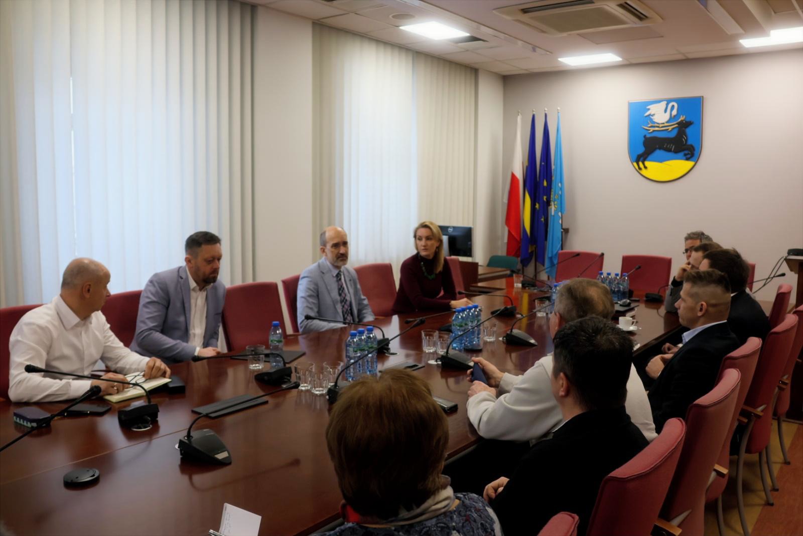 Osoby siedzące za stołem konferencyjnym w pomieszczeniu. Prowadzone są rozmowy 