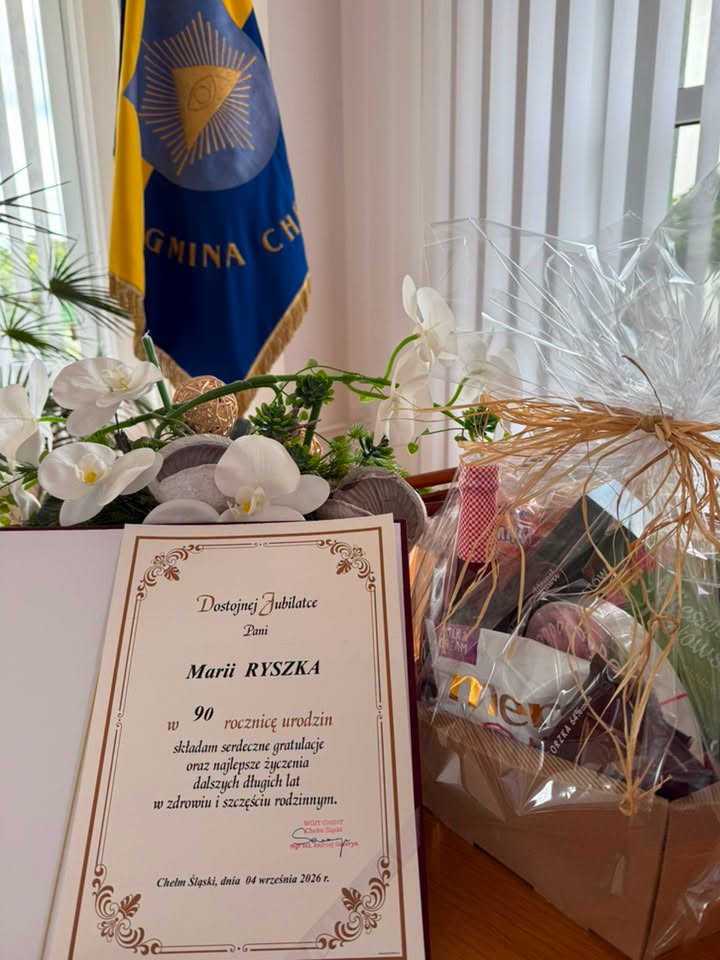Życzenia dla Jubilatki, na stole kwiaty i sztandar Gminy w oddali 