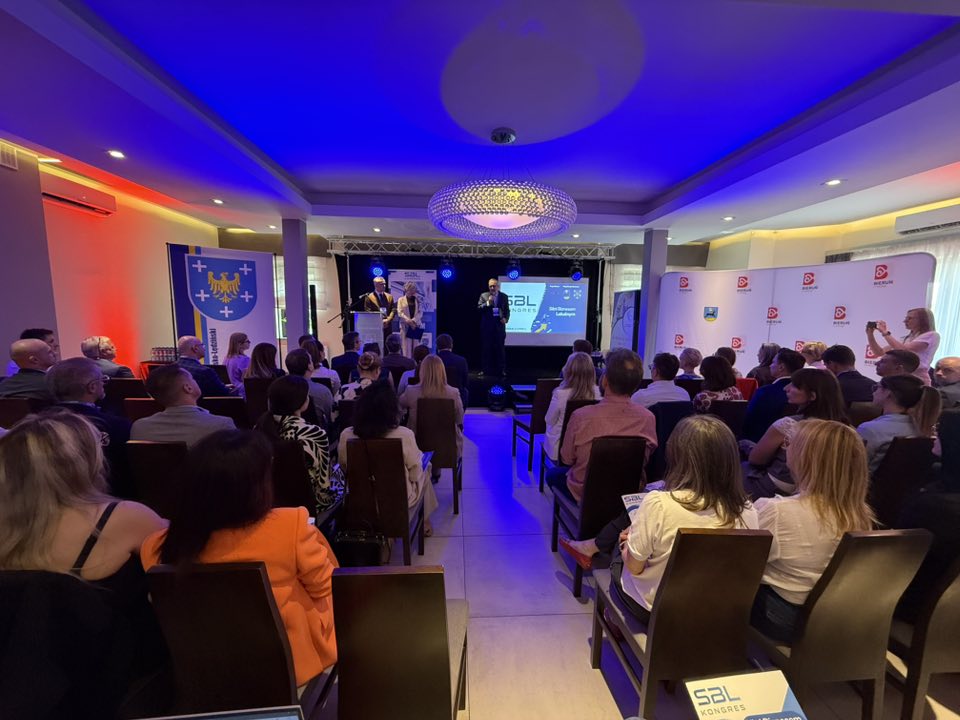 Sala konferencyjna wypełniona uczestnikami. Siedzą na krzesłach przed sceną. Na scenie są mówcy. 