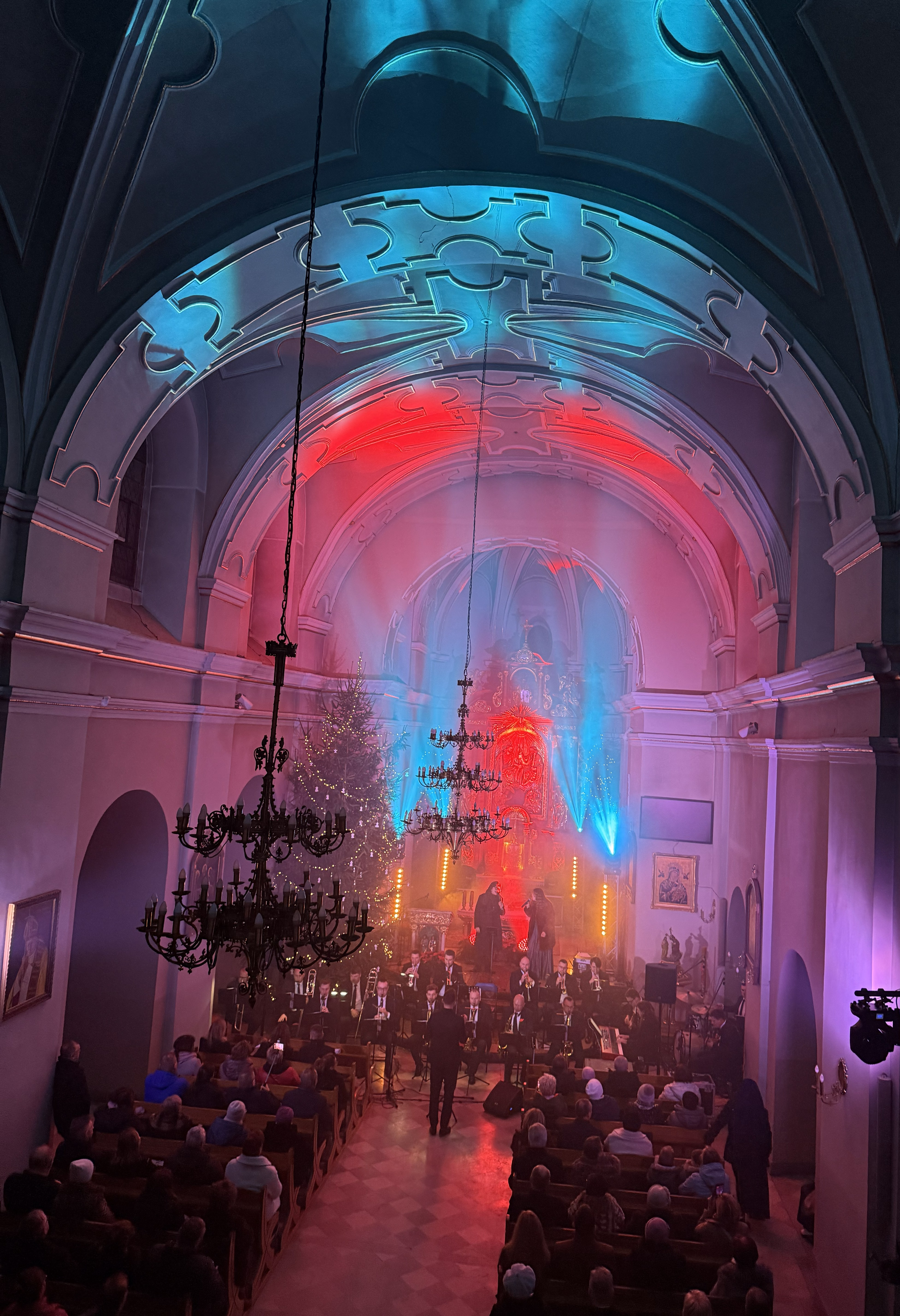 Wnętrze kościoła oświetlone kolorowym światłwem , przed ołtarzem zasiadający muzycy i śpiewający wokaliści. Wierni siedzący w ławkach po prawej i lewej stronie 