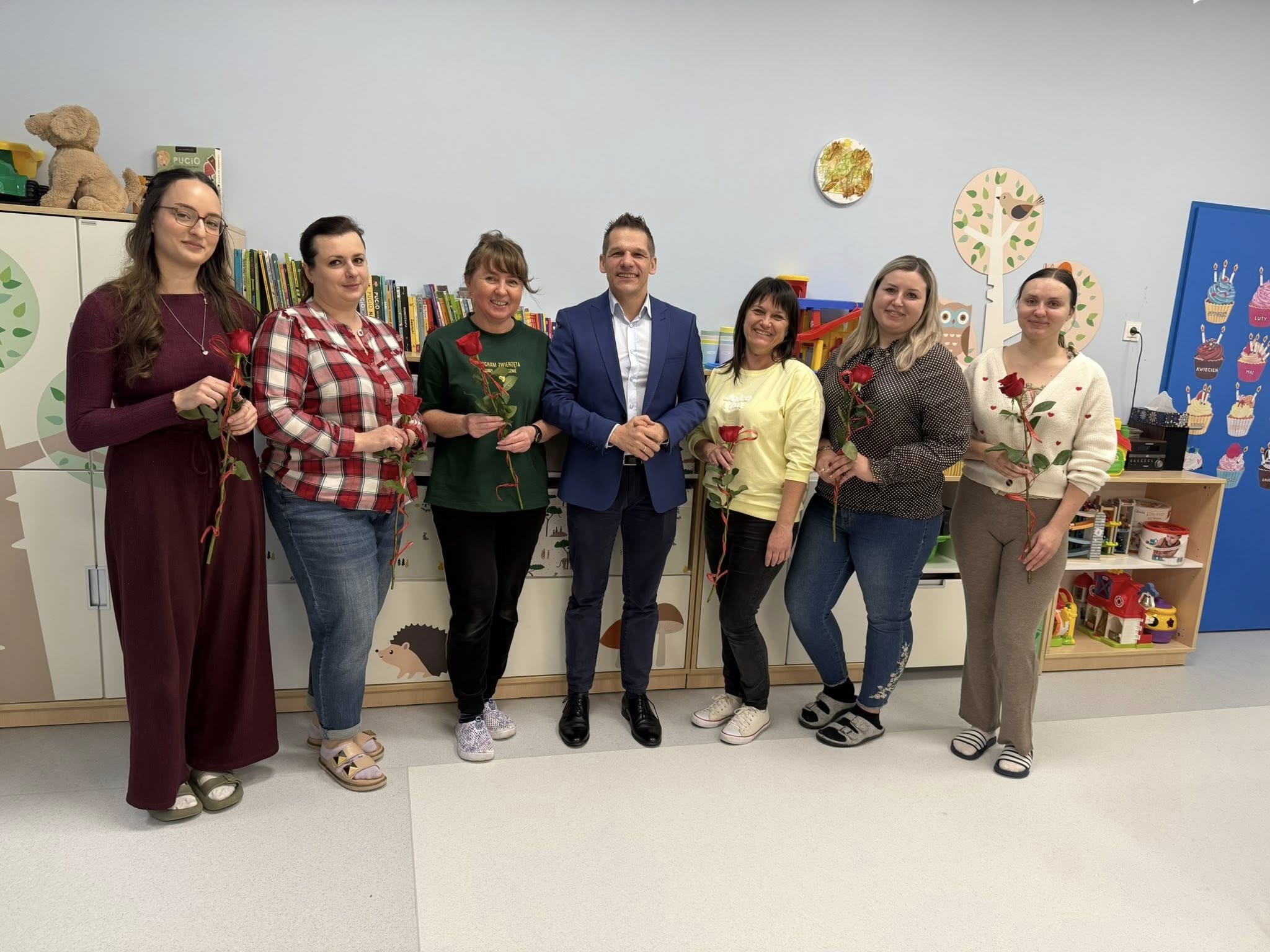 Sześć kobiet i mężczyzna pozujący do zdjęcia. Kobiety trzymają w ręce róże. Wszyscy stoją w sali zajęć na tle kolorowych, dziecięcych mebli.