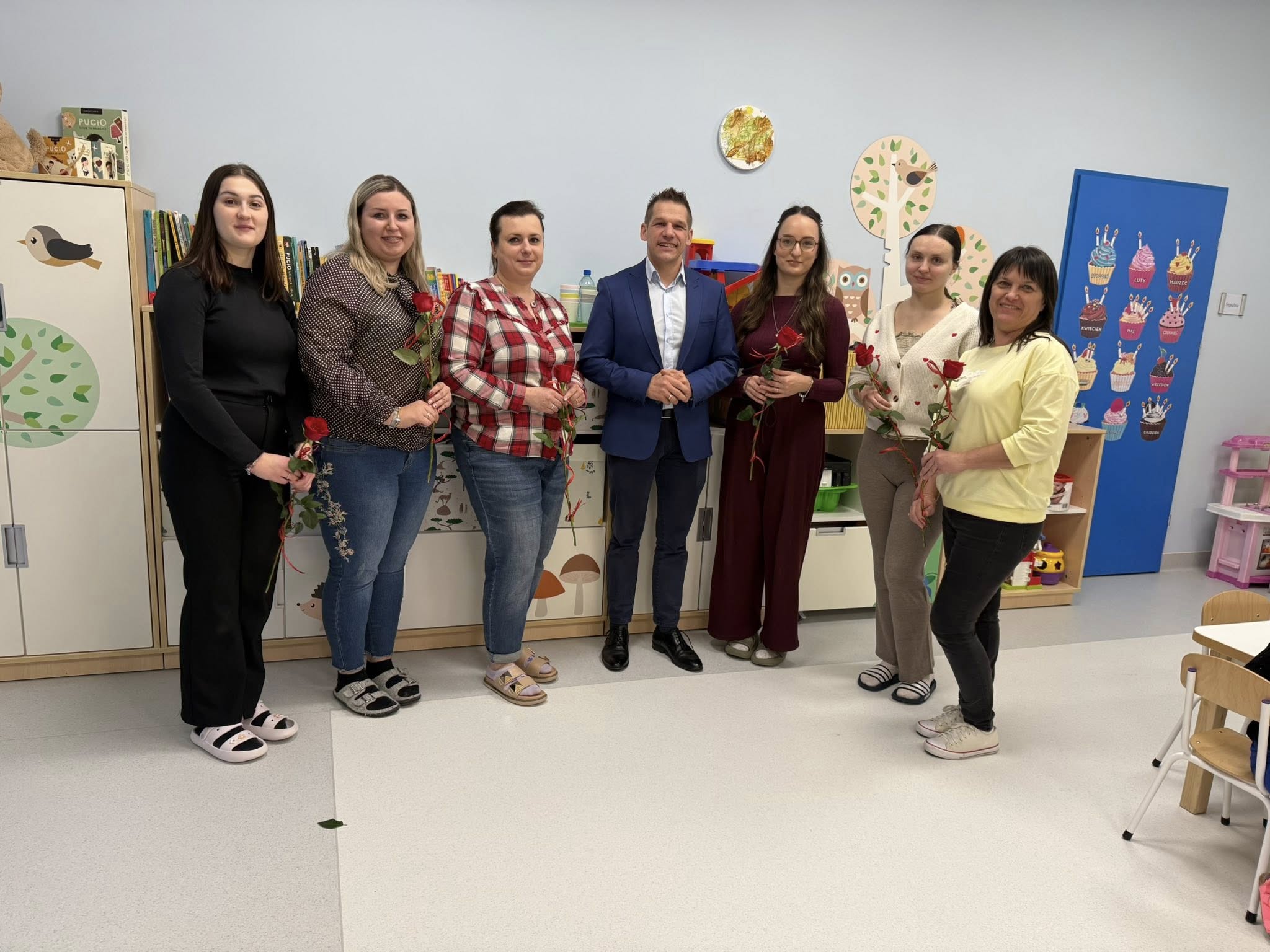 Sześć kobiet i mężczyzna pozujący do zdjęcia. Kobiety trzymają w ręce róże. Wszyscy stoją w sali zajęć na tle kolorowych, dziecięcych mebli i zabawek. 