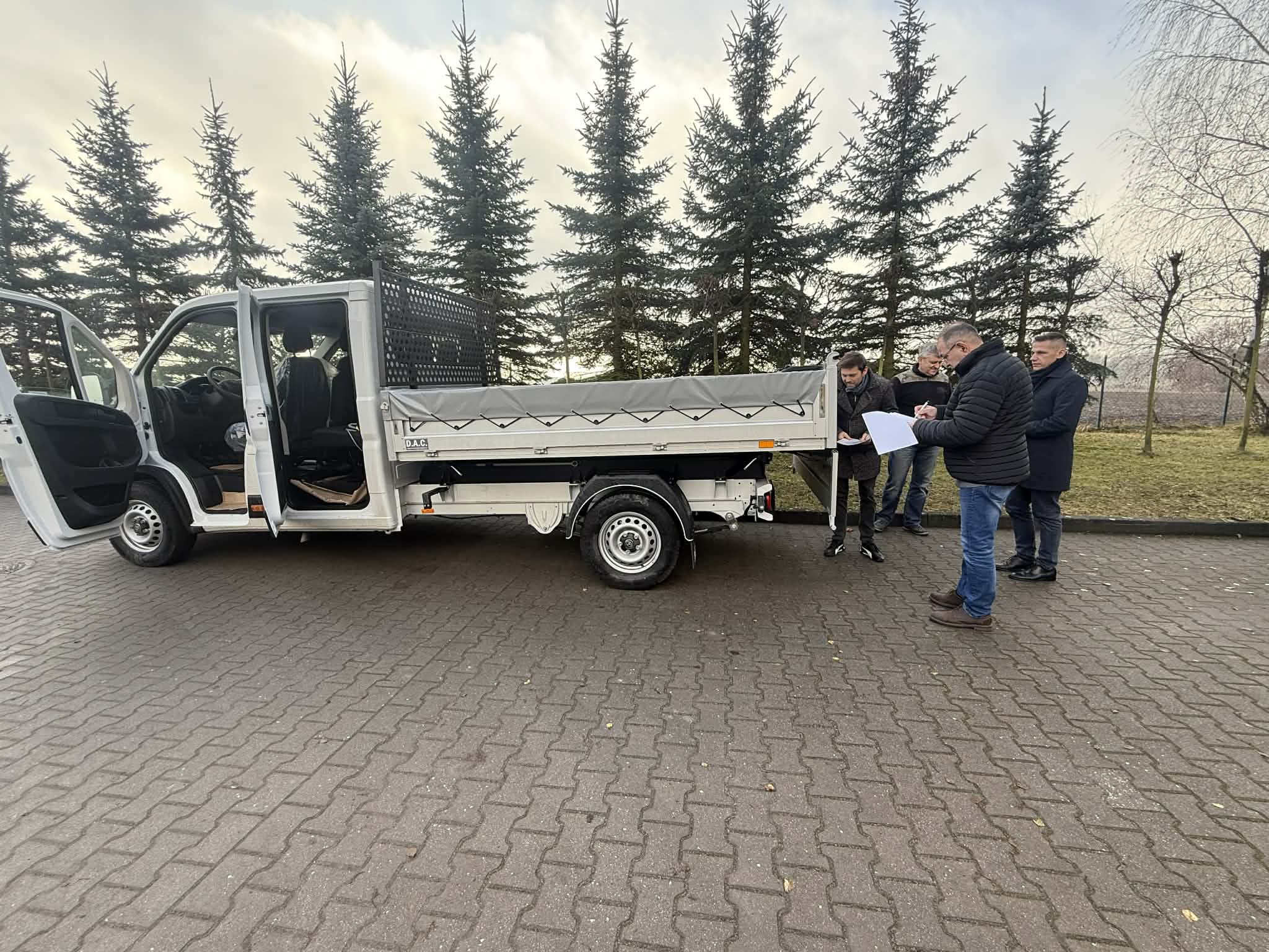 Samochód dostawczy z paką i plandeką- widok z boku. Panowie dokonujący odbioru. 