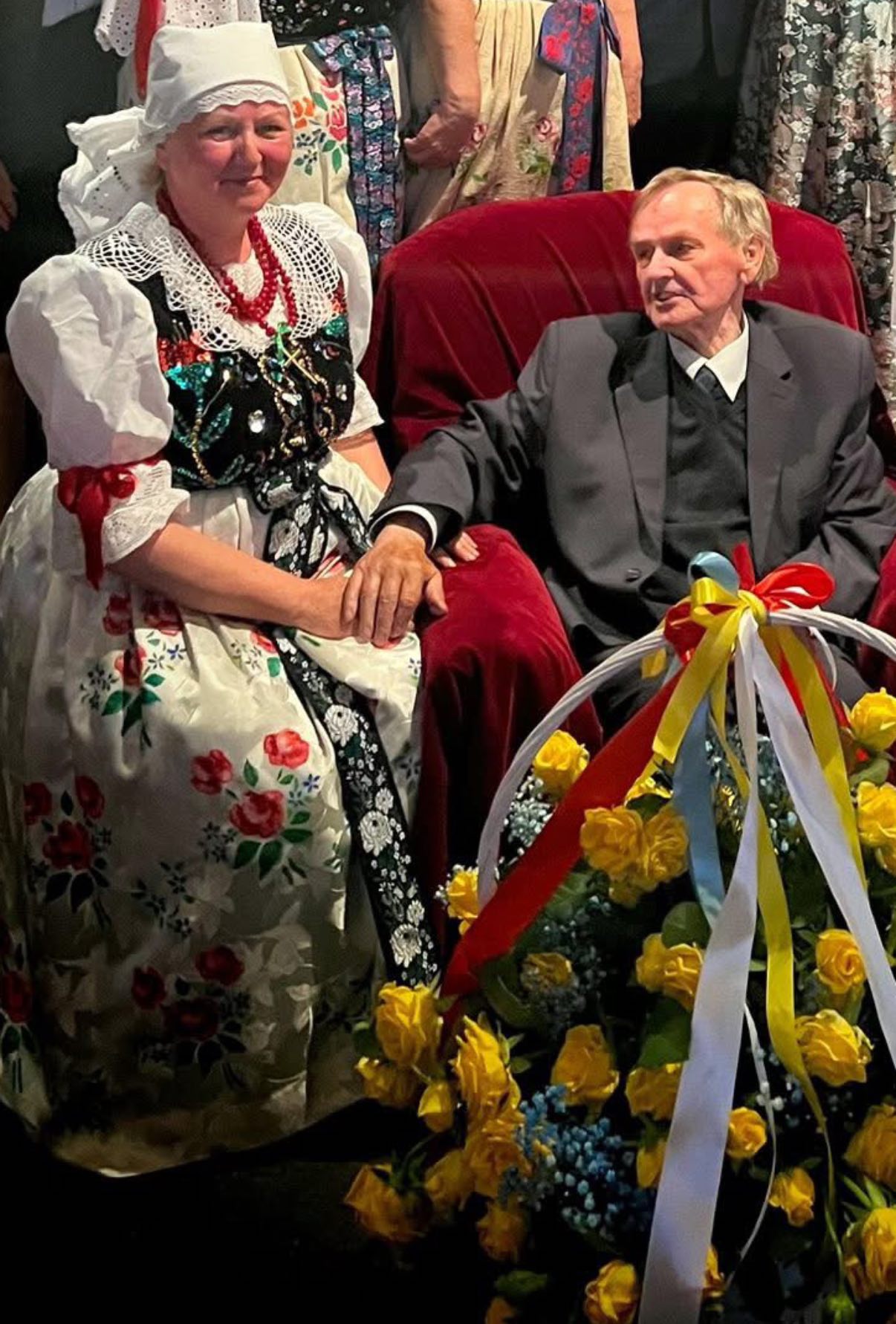 Kobieta w stroju ludowym, mężczyzna siedzący w fotelu z koszem kwiatów stojacym obok 
