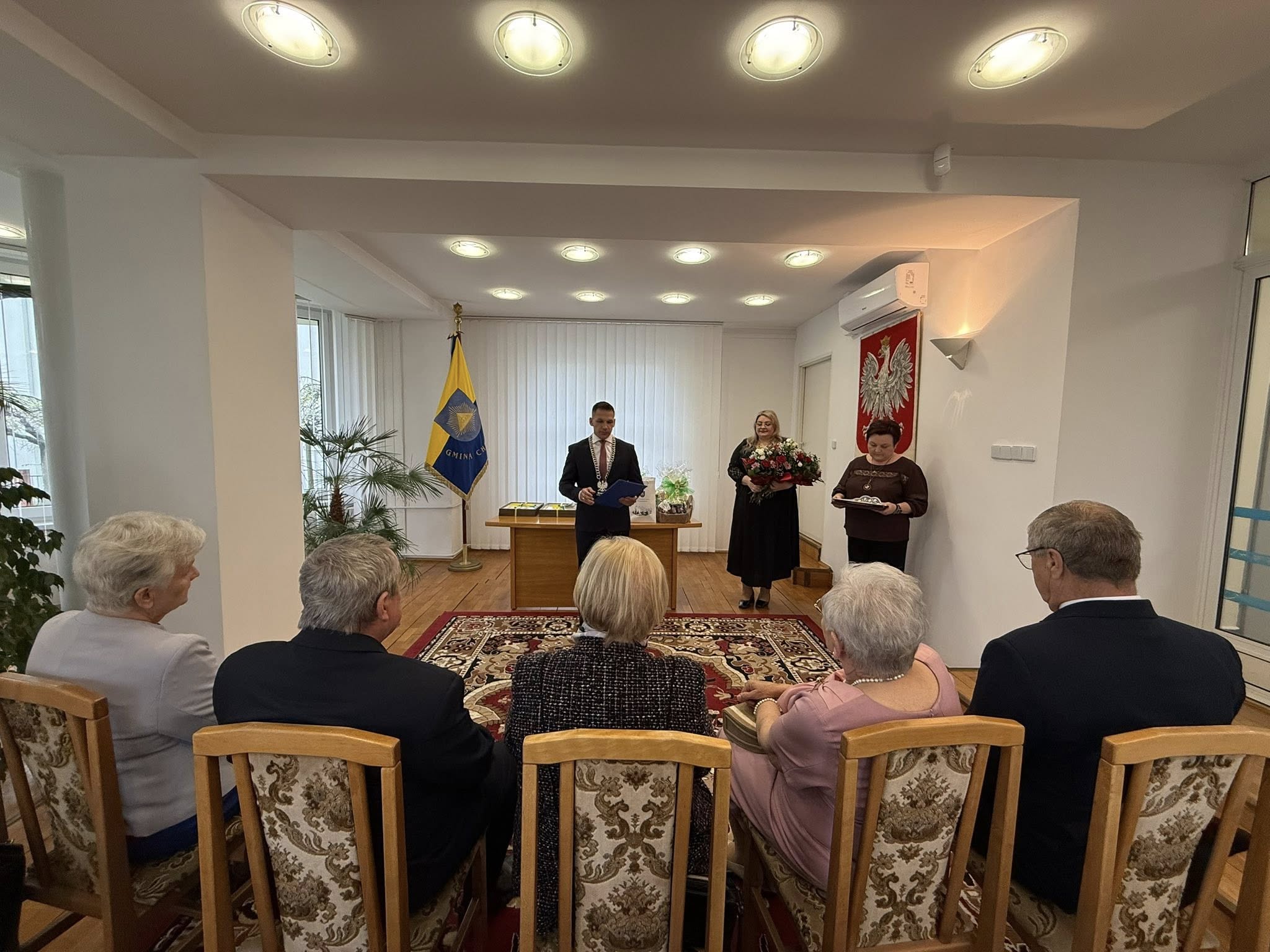 Osoby w pomieszczeniu. Pięc osób siedzi na krzesłach a trzy stoją zwrócone do nich twarza. 