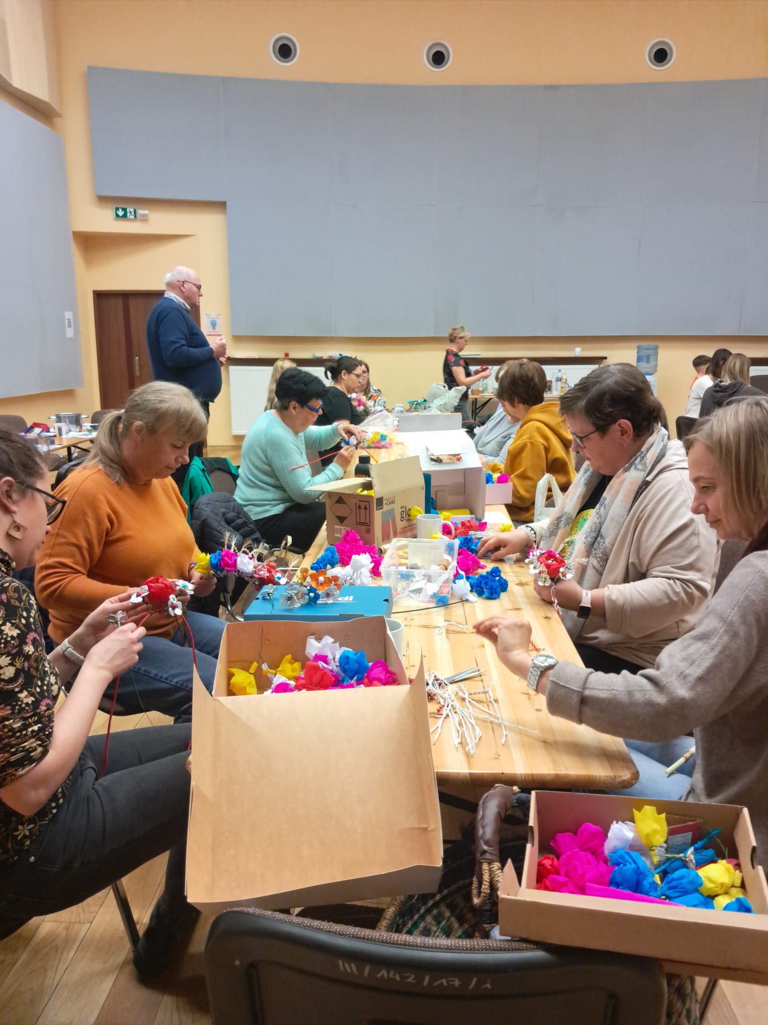 Osoby sidzące i stojące w pomieszczeniu przy stołach na warsztatach tworzące kwiaty , ozdoby do glandy 