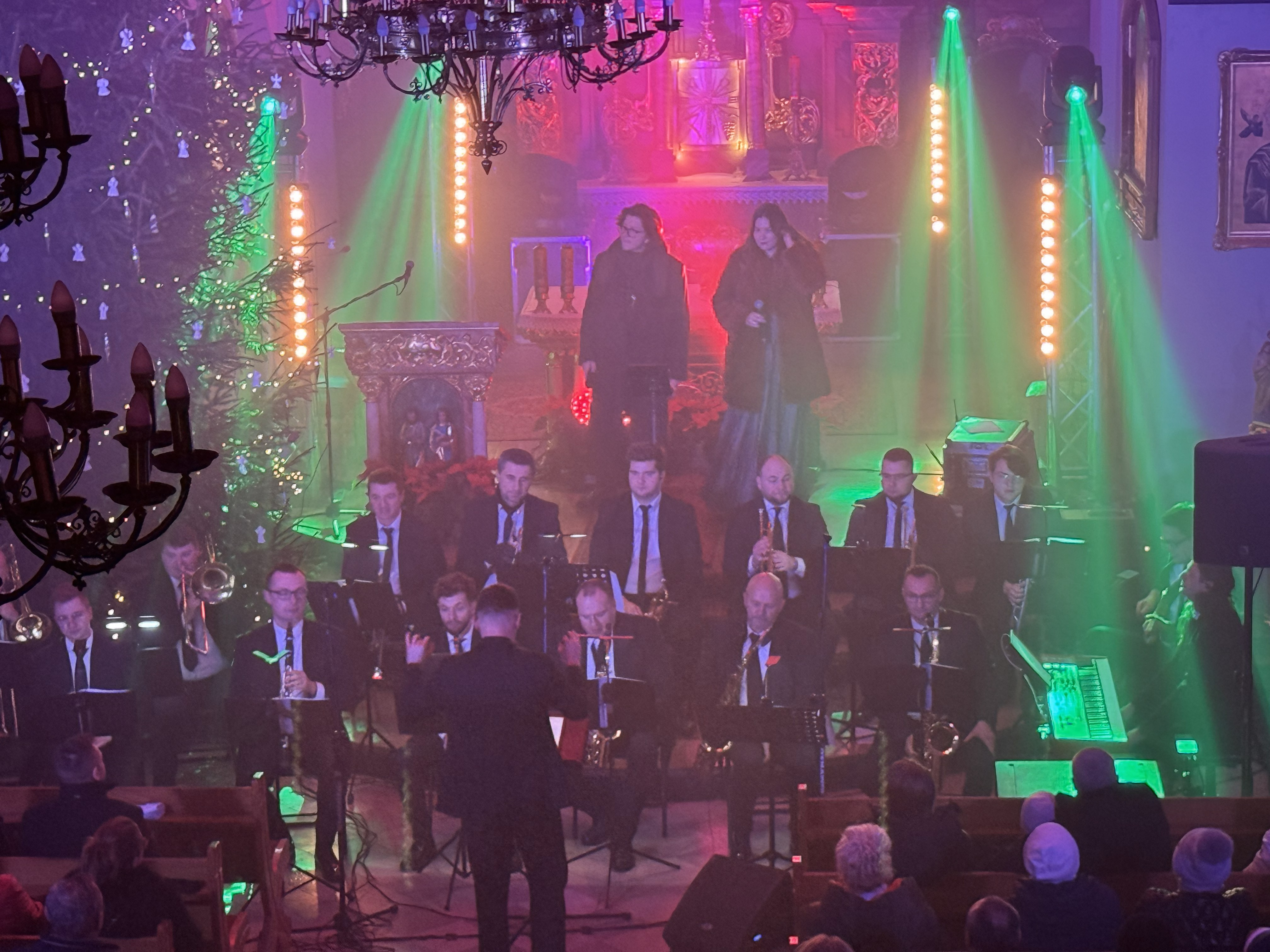 Wnętrze kościoła oświetlone kolorowym światłwem , przed ołtarzem zasiadający muzycy i śpiewający wokaliści. Wierni siedzący w ławkach po prawej i lewej stronie 