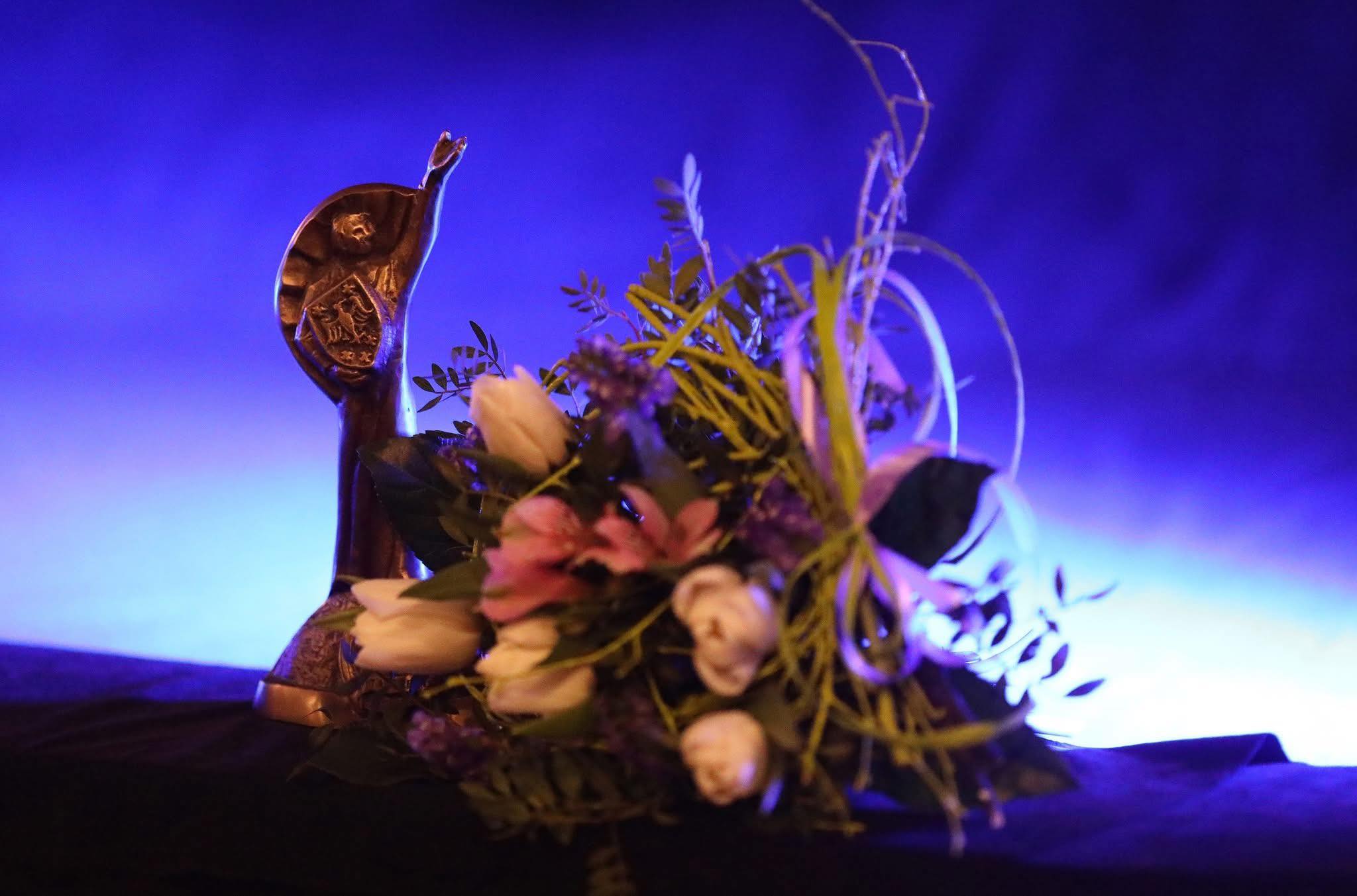 Na granatowym tle ustawiona jest statuetka metalowego Clemensa i bukiet kwiatów 