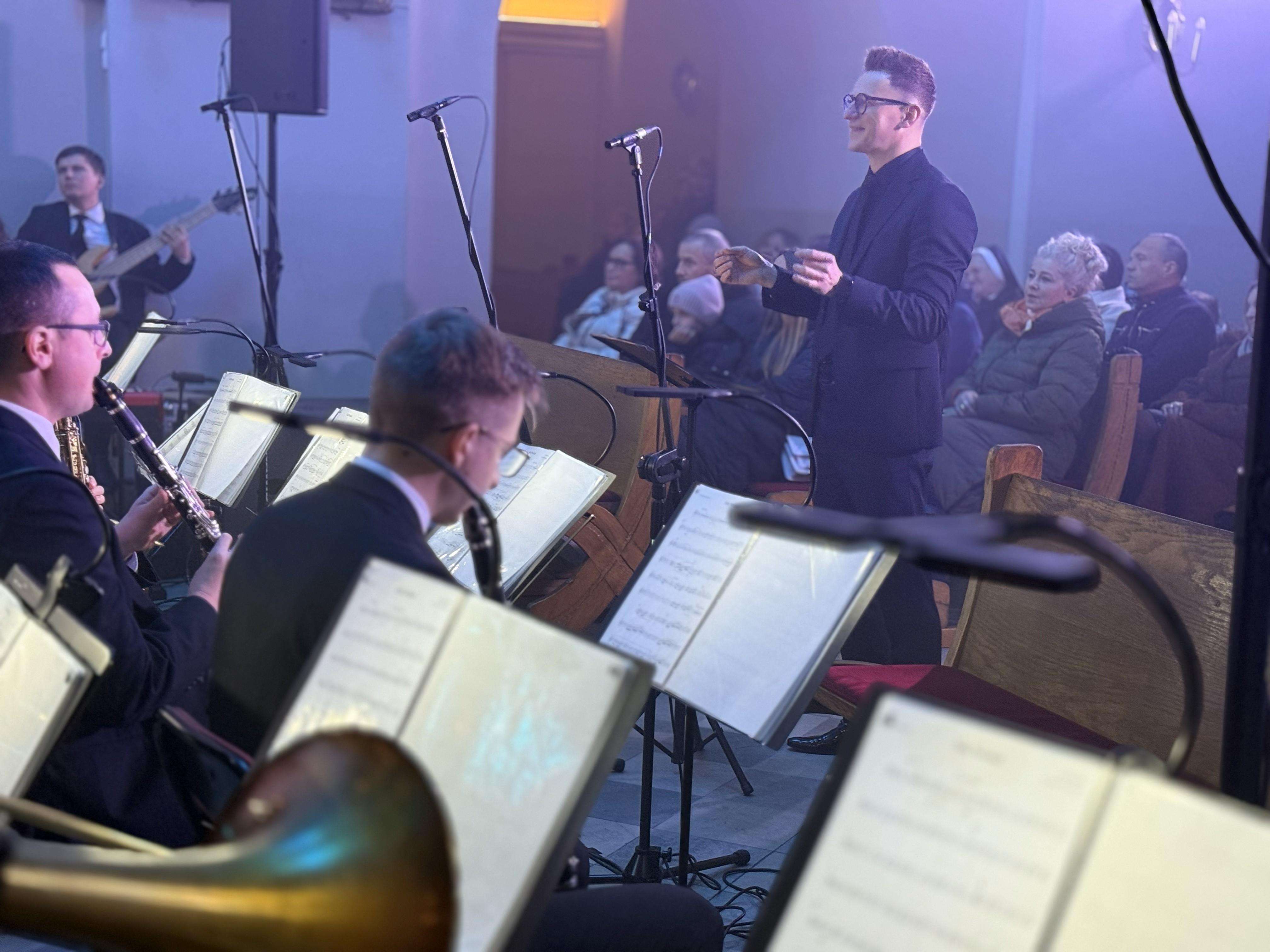 Wnętrze kościoła oświetlone kolorowym światłwem , przed ołtarzem zasiadający muzycy i dyrygujący mężczyzna. Wierni siedzący w ławkach po prawej i lewej stronie 