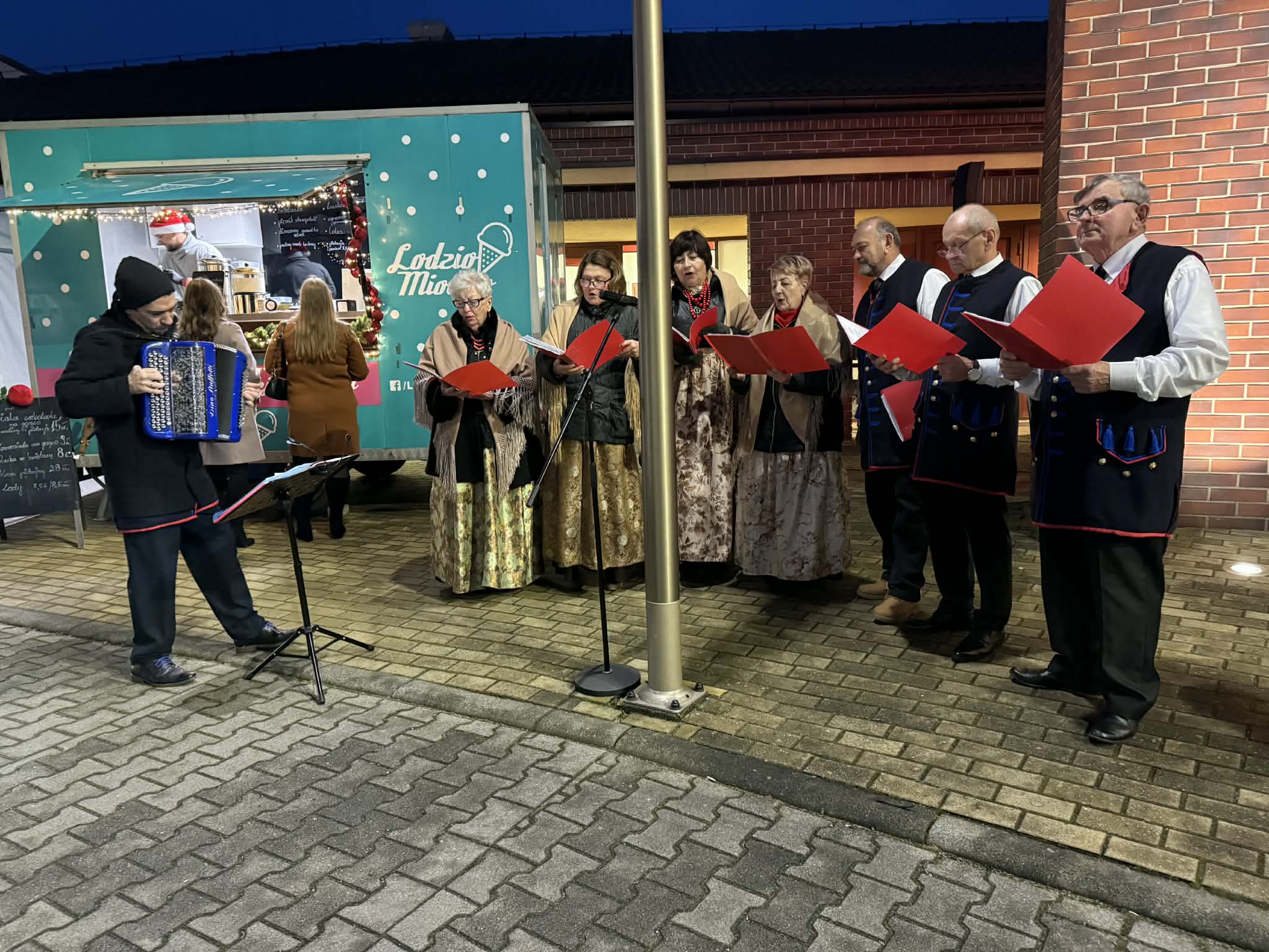 Mężczyżni i kobiety śpiewające w strojach ludowych. Mężczyzna gra na akordeonie. W tle stoisko gastronomiczne 