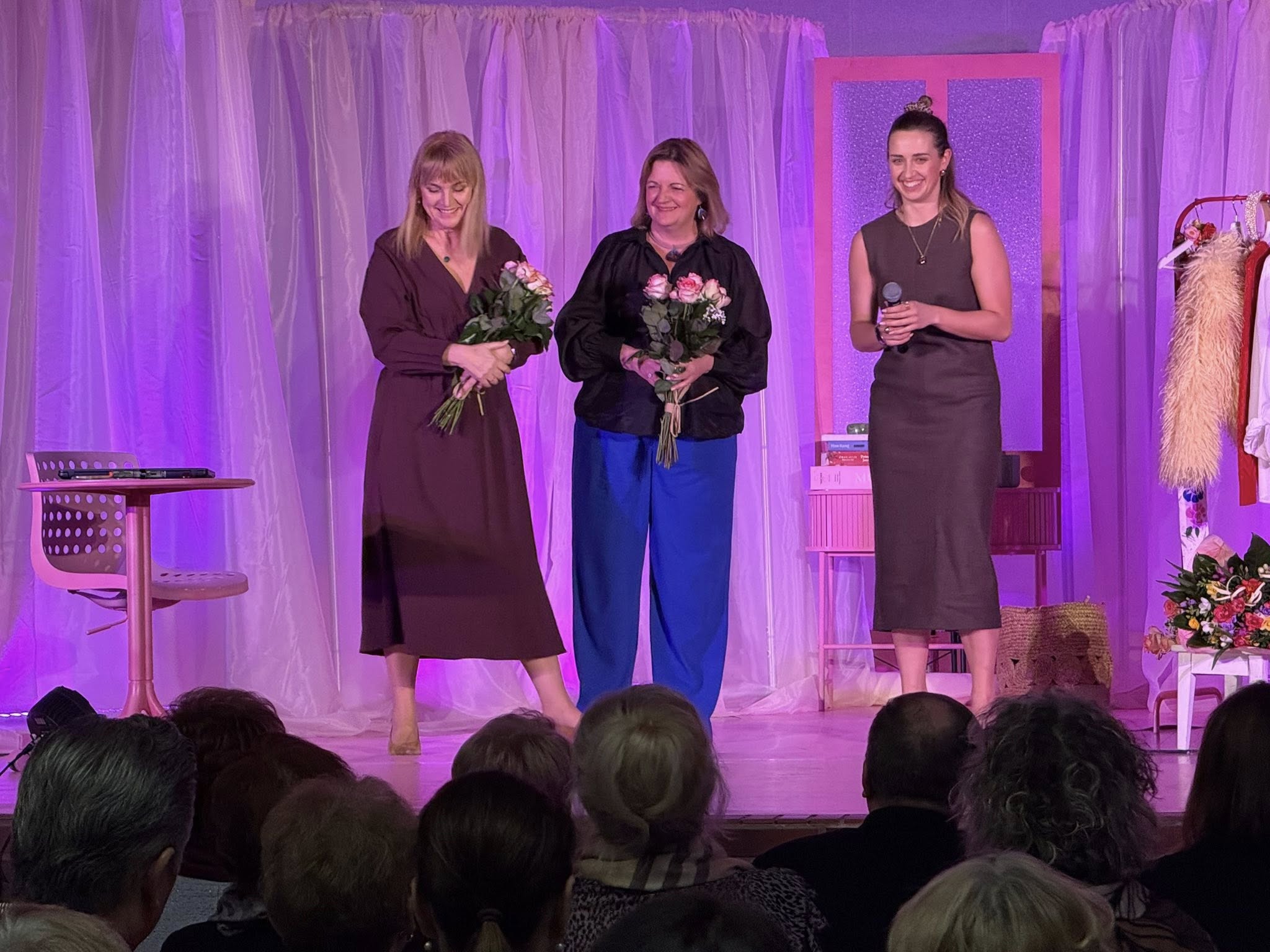 Trzy kobiety stojące na scenie. Trzymają w rękach kwiaty 