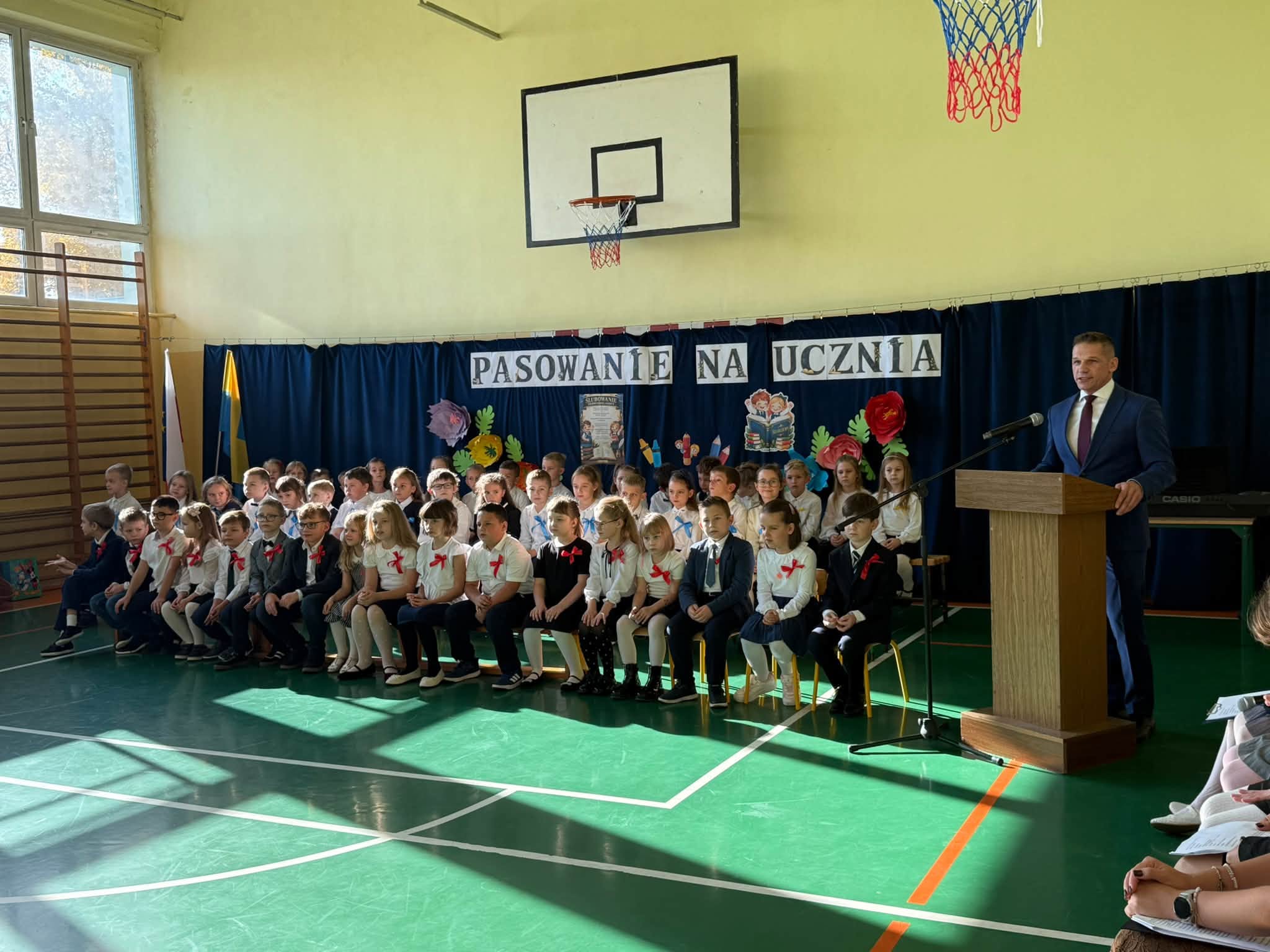 Uczniowie siedzacy w sali gimnastycznej podczas akademii i przemawiajacy mężczyzna przy mównicy