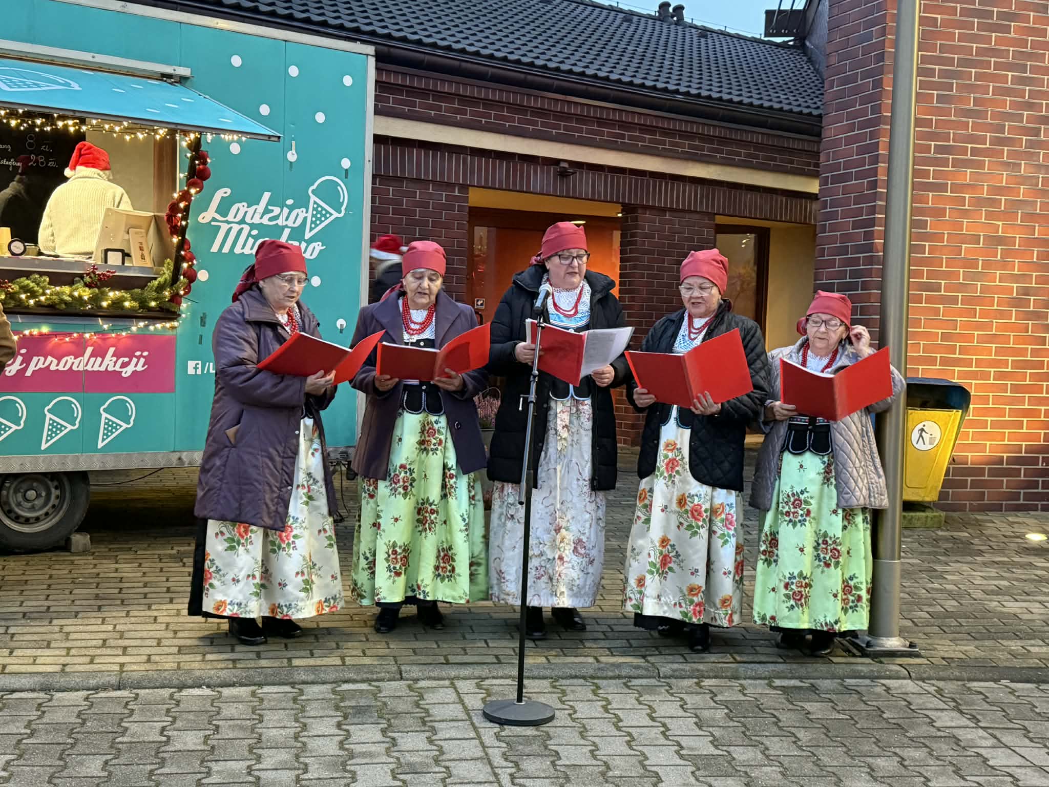 Kobiety stojące przed budynkiem w strojach ludowych i śpiewające. 
