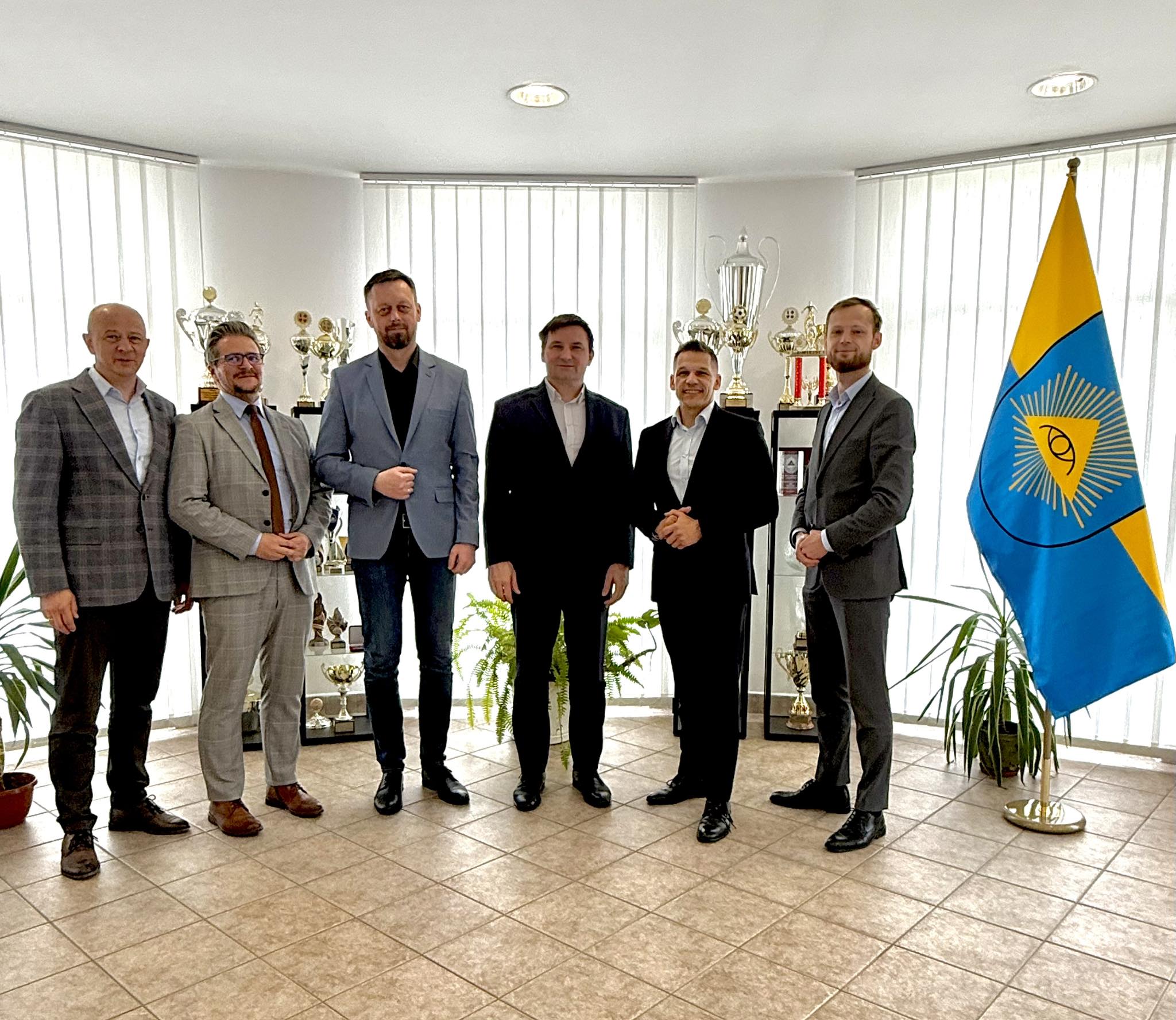 Mężczyźni pozujący do zdjęcia w pomieszczeniu . Obok stoi flaga Chełmu Ślaskiego 