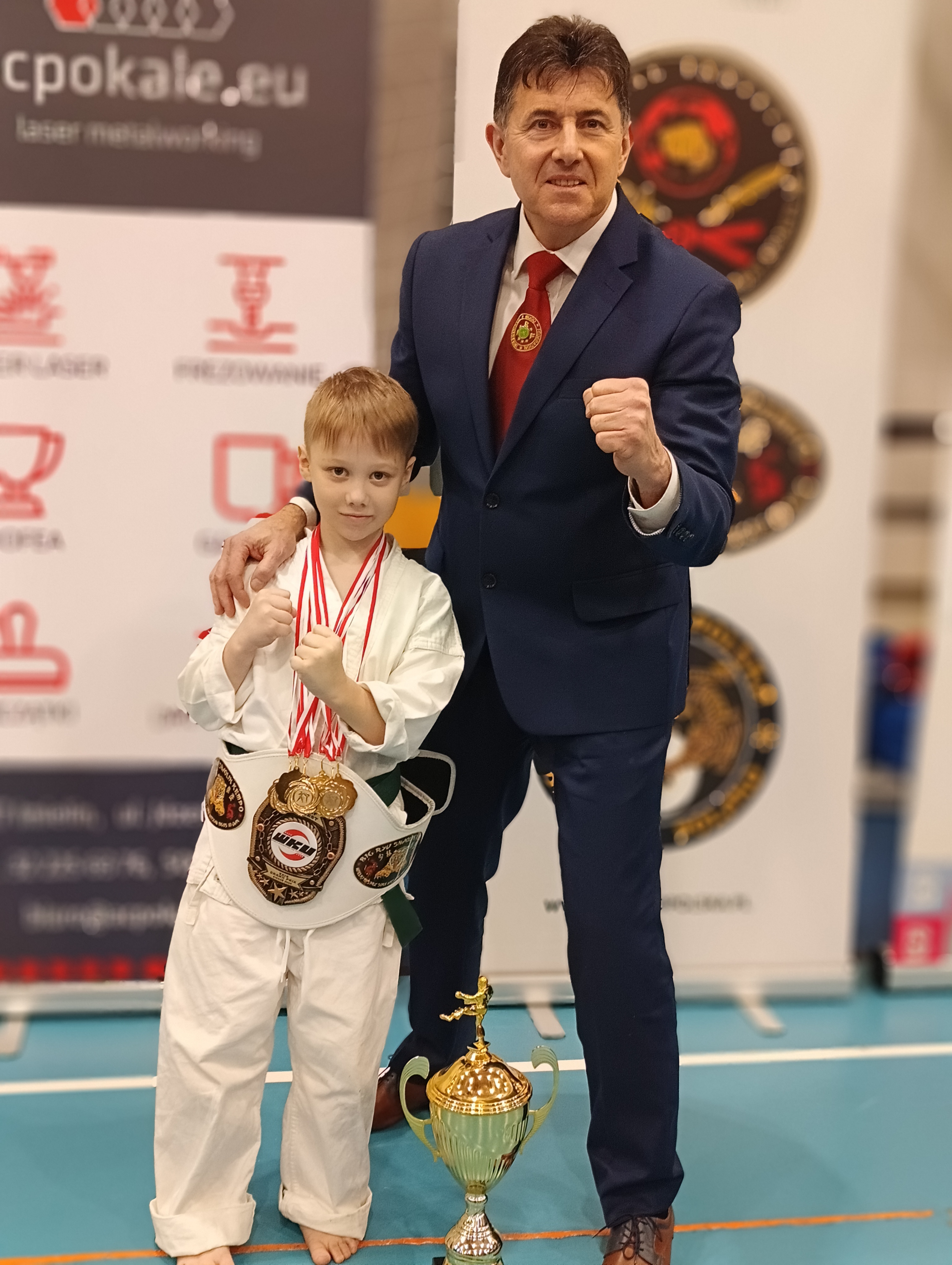 Młody zawodnik ubrany w kimono i przyozdobiny medalmi, a także pasem mistrza. Z chłopcem na zdjęciu mężczyzna - trener. W tle hala sportowa. 