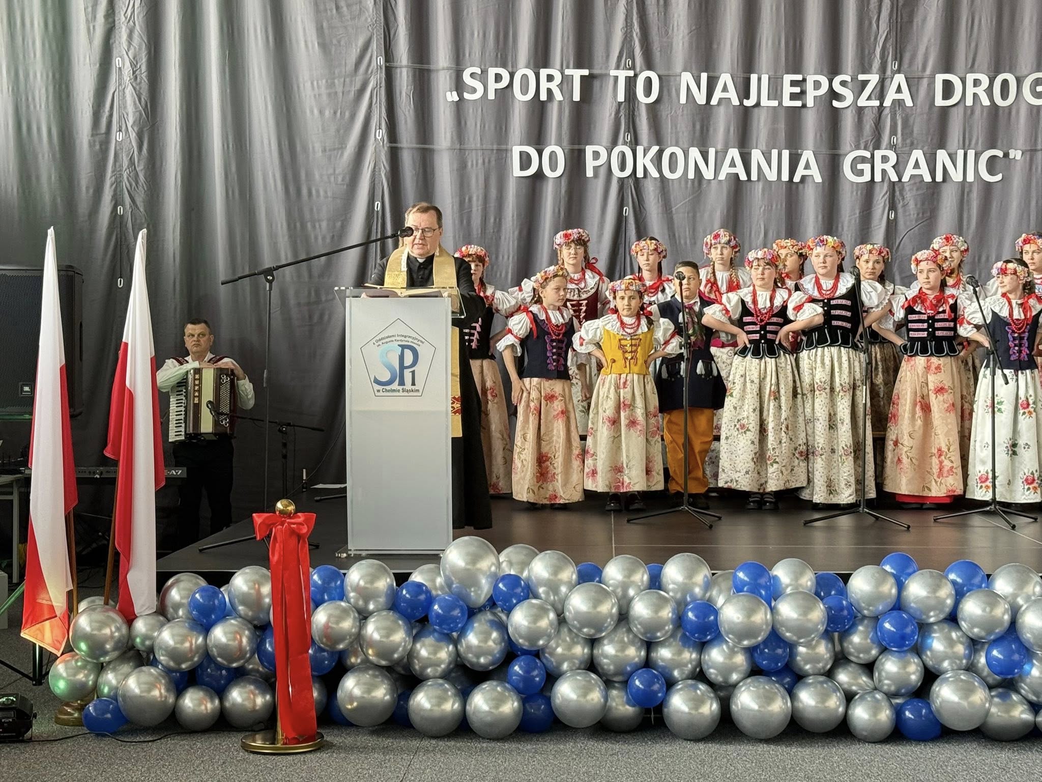 Dzieci w strojach ludowych na scenie, a przed nimi przy mówinicy ksiądz 