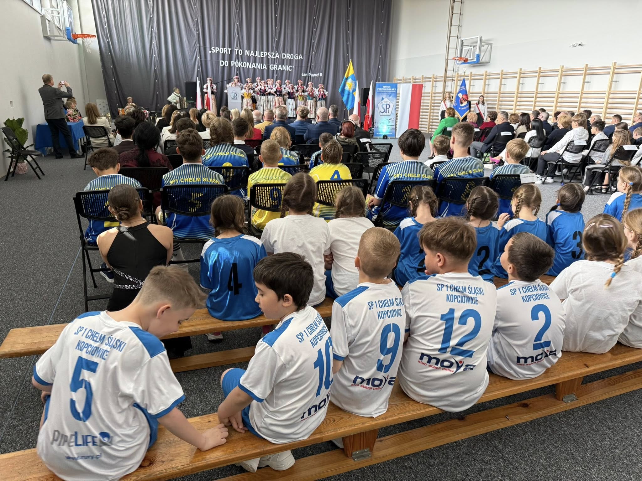 Osoby siedzące w pomieszczeniu sali gimnastycznej. Przed nimi scena i osoby występujące. 