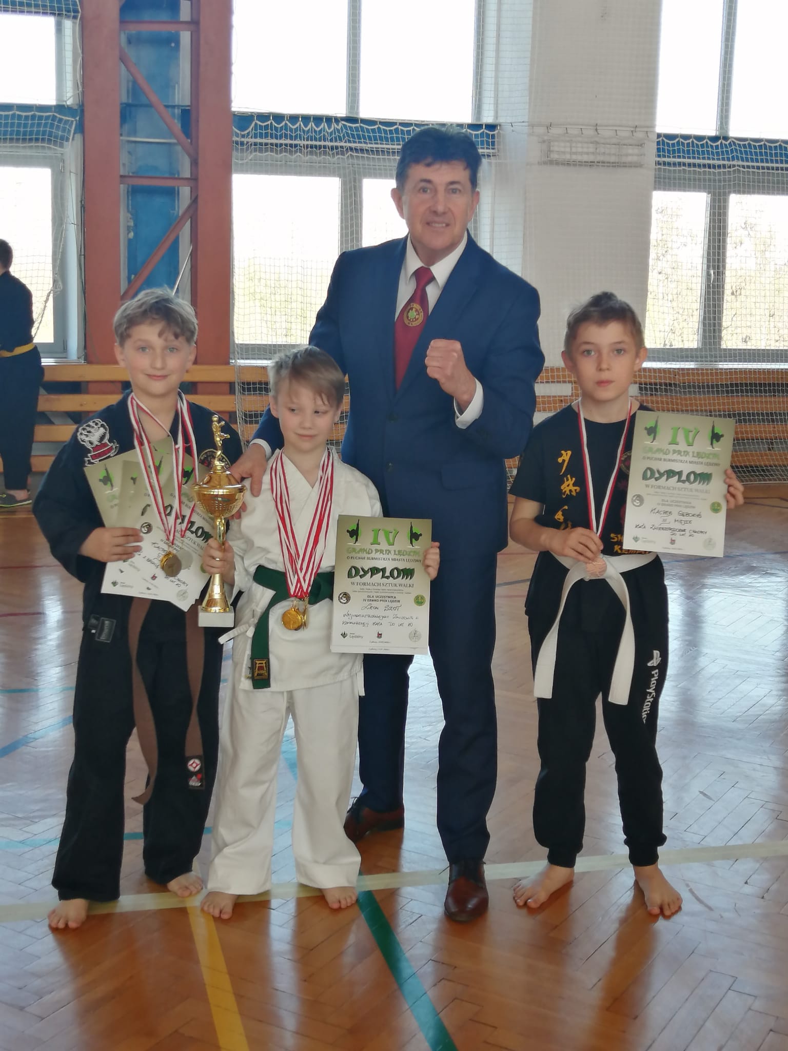 Trzej chłopcy w strojach sportowych i mistrz trzymający w dłoniach dyplomy i medale 