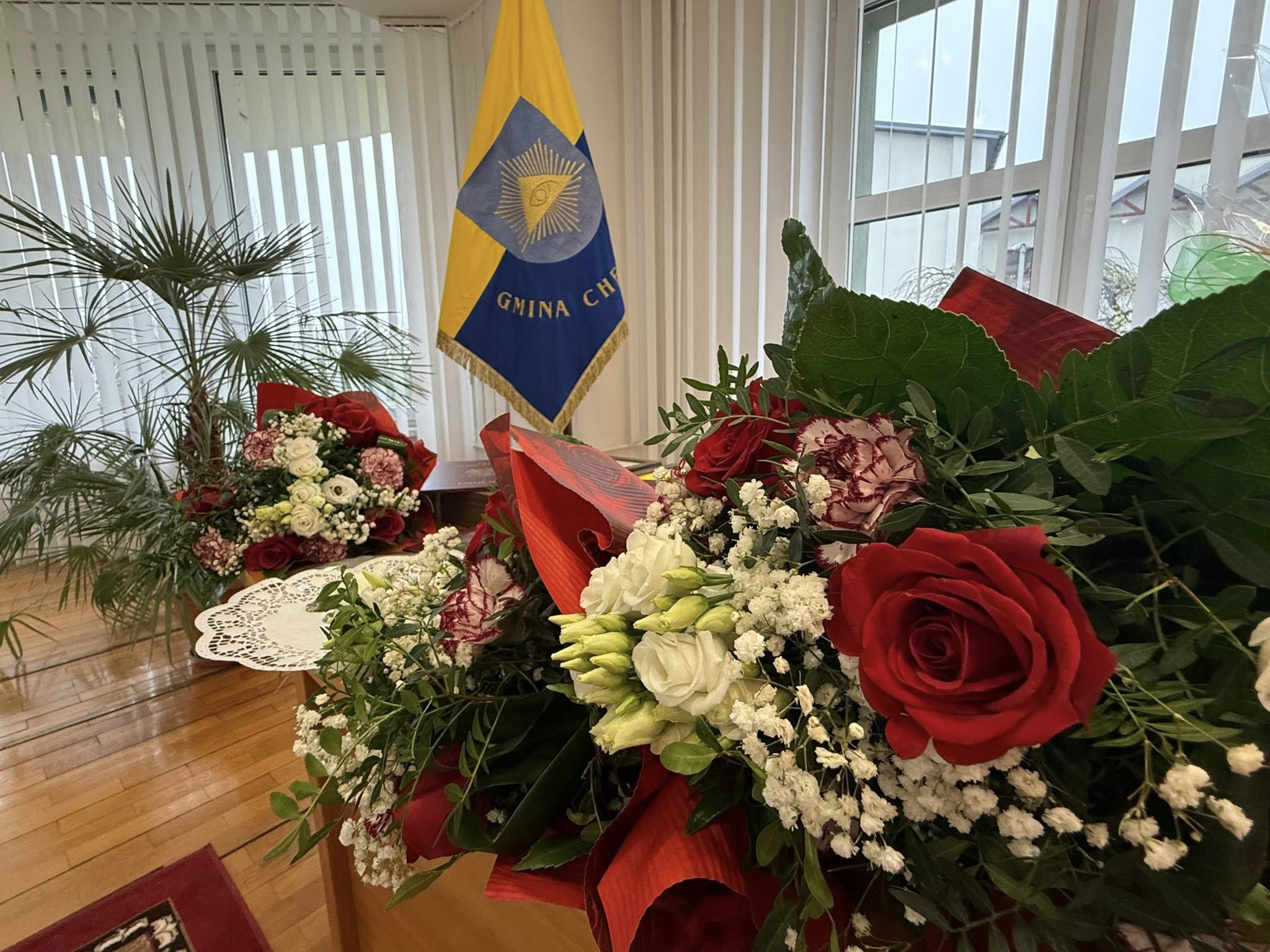 Kiaty na stoliku w pomieszczeniu - bukiet z róż, sztandar gminy i kwiaty. 