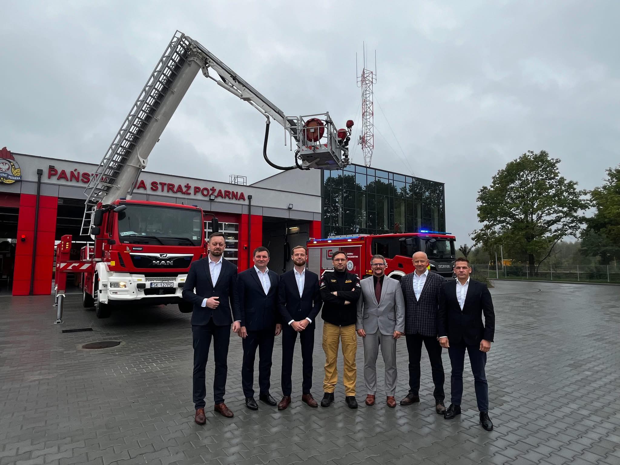 7 mężczyzn pozujących do zdjęcia przed jednostką JRG. W tle budynek jednoski i wóz specjalistyczny. 
