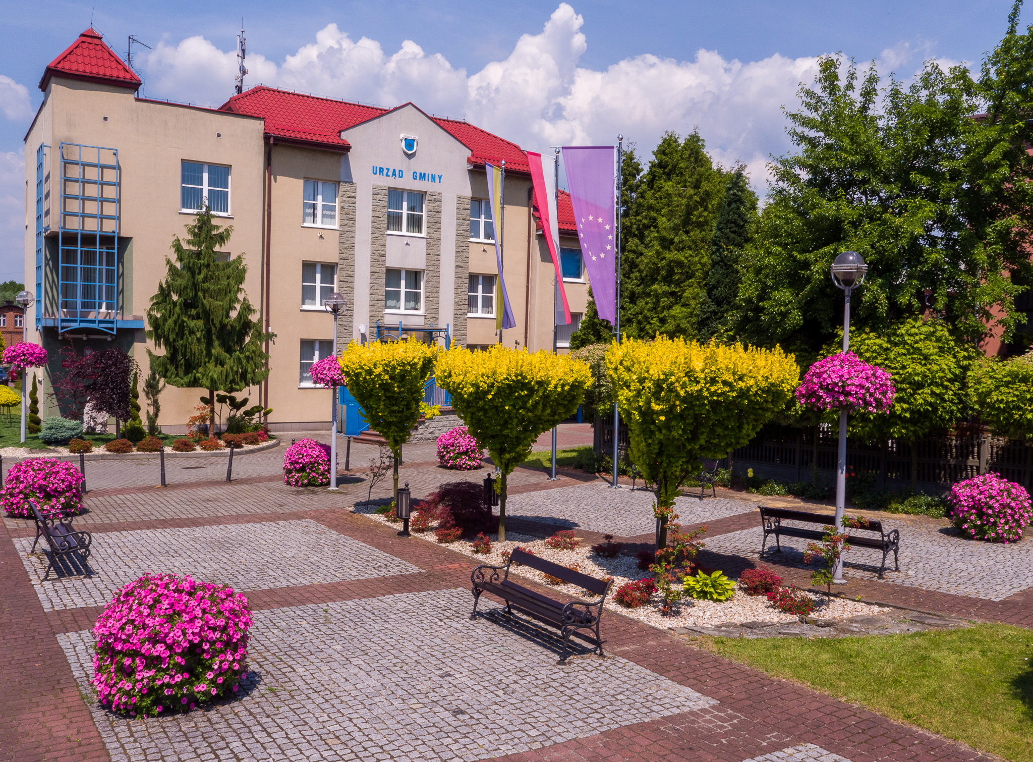 Budynek urzędu z rabatami kwiatowyi przed budynkiem, flagami. Ściana frontowa obiektu. 
