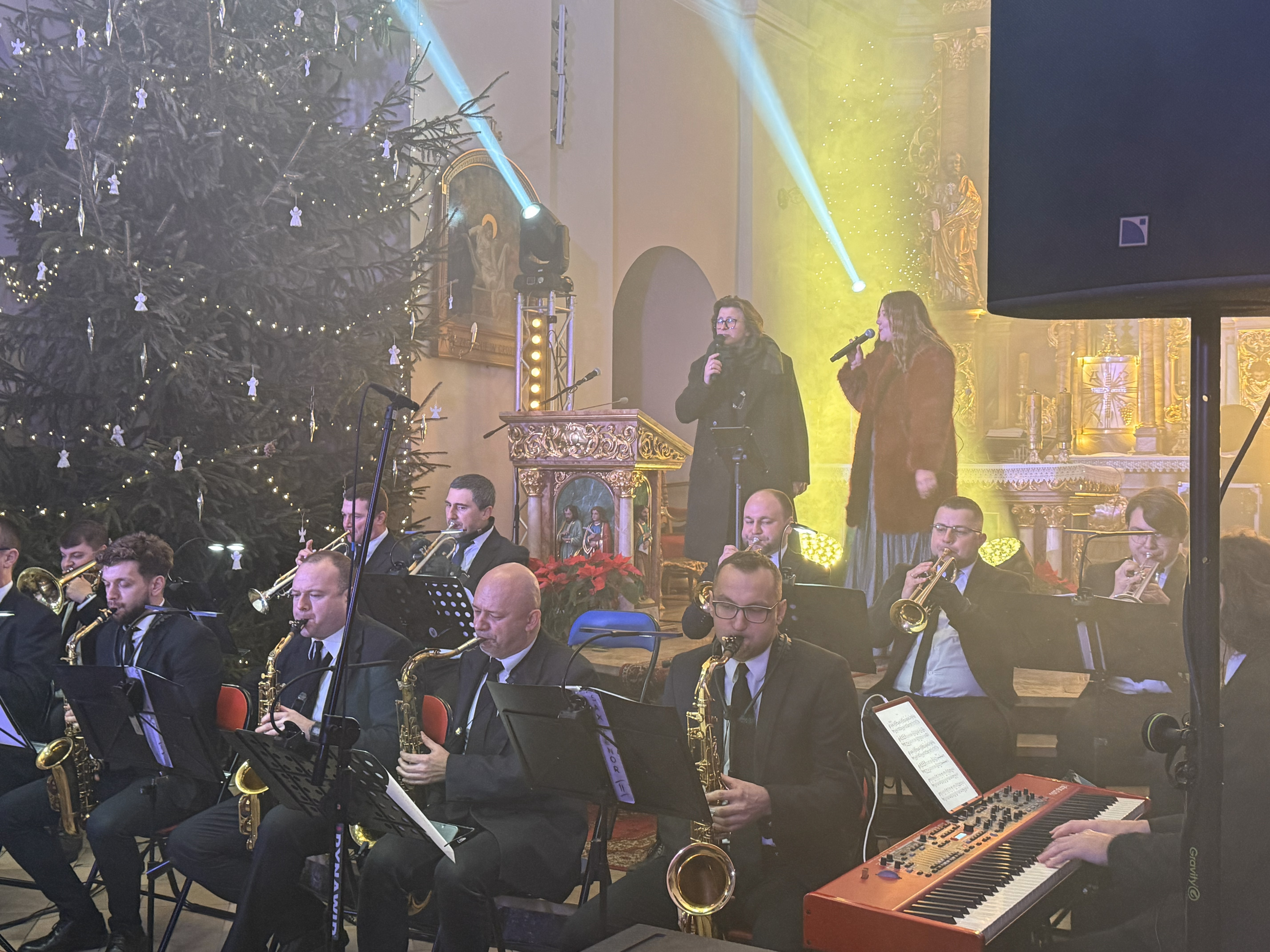 Wnętrze kościoła oświetlone kolorowym światłwem , przed ołtarzem zasiadający muzycy i śpiewający wokaliści. 