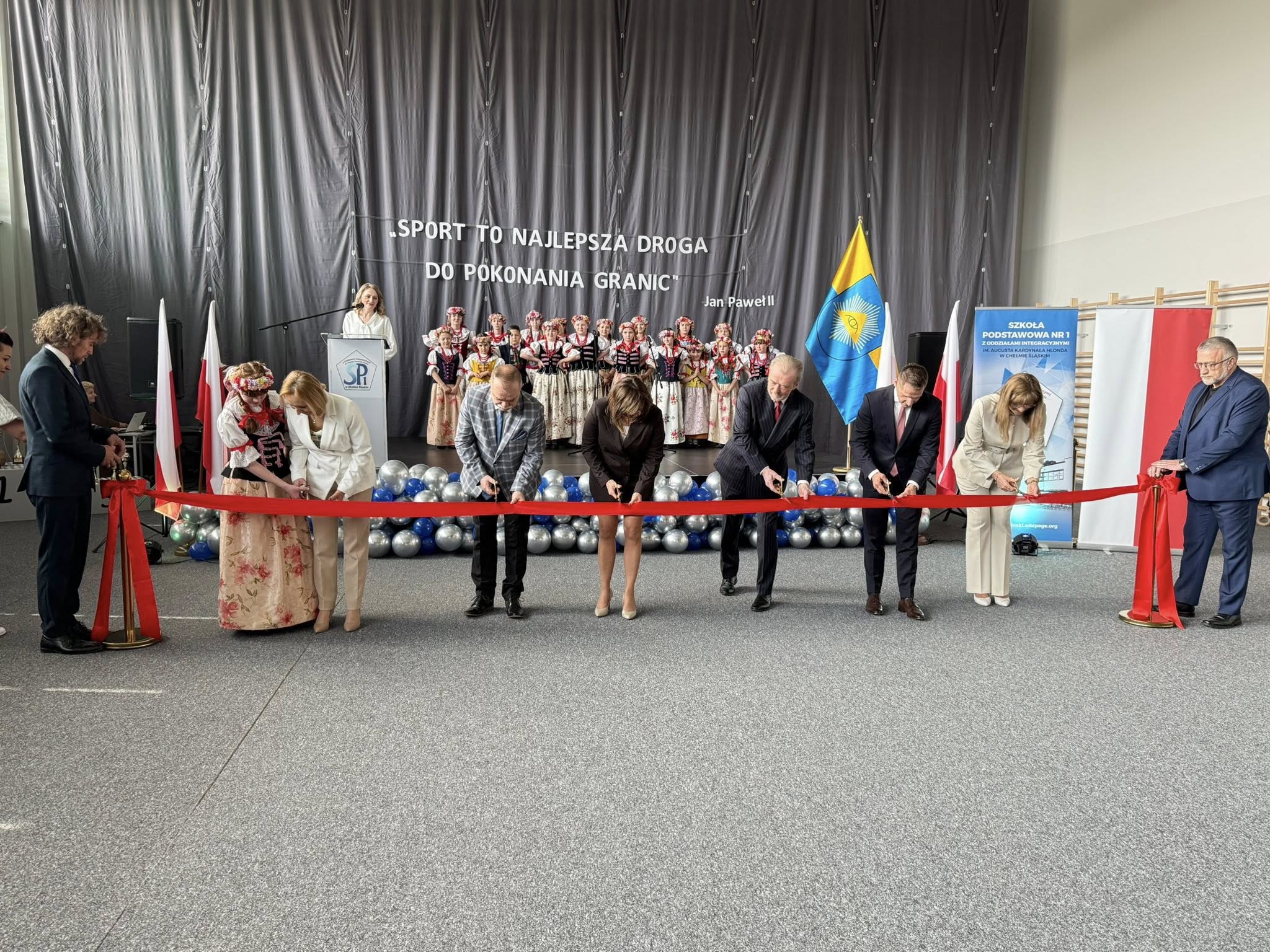 Osoby przecinające wstęgę. Za nimi na scenie młodzież w strojach ludowych . 