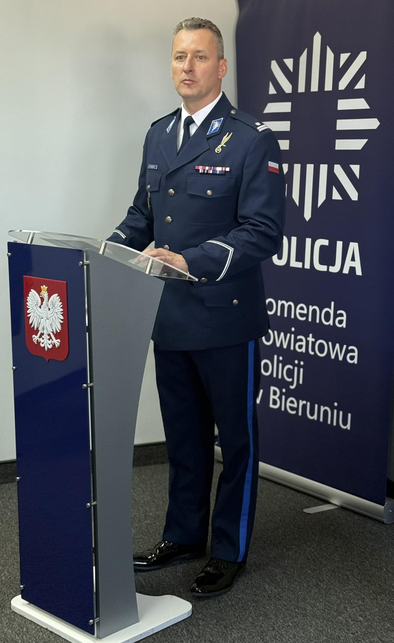 Mężczyzna w mundurze policyjnym stojący za mównicą w pomieszczeniu 